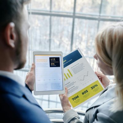 Two business professionals reviewing graphs and data on a tablet and paper report indoors.