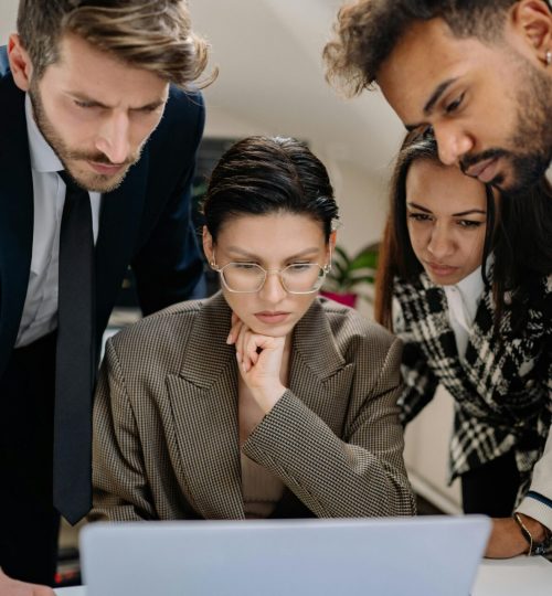 A diverse group of professionals intensely collaborating over a laptop indoors.